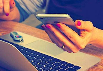 Frau sitzt vor Laptop und hält Smartphone in ihrer linken Hand