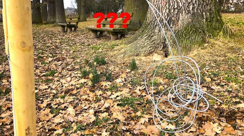 Sabotierter Zaun im Eichenhain vor Sitzbank mit drei roten fotomontierten Fragezeichen