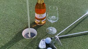 Sektflasche, Golfschläger und -bälle neben einem Loch auf einem Putting Green