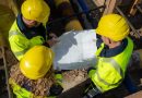 Drei Mitarbeiter mit gelben Warnwesten und Bauhelmen betrachten in einer Baugrube einen Plan