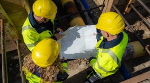 Drei Mitarbeiter mit gelben Warnwesten und Bauhelmen betrachten in einer Baugrube einen Plan
