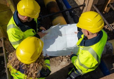 Drei Mitarbeiter mit gelben Warnwesten und Bauhelmen betrachten in einer Baugrube einen Plan